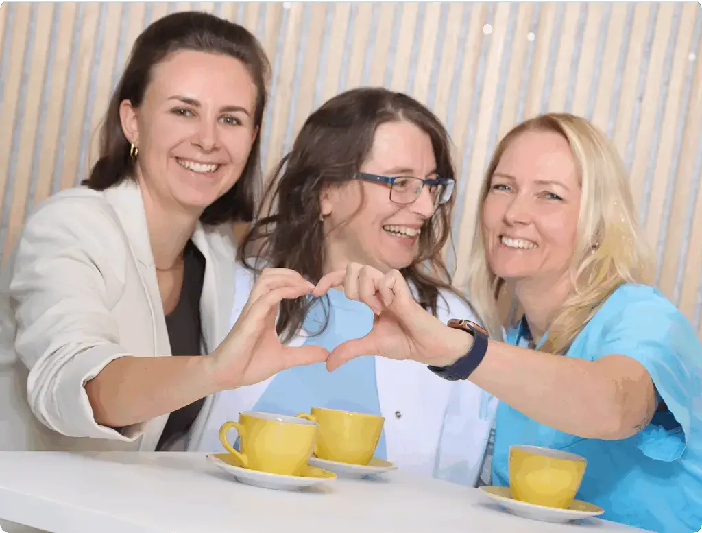 erinnen sitzen lachend zusammen, zwei von ihnen formen mit den Händen ein Herz, vor ihnen stehen Kaffeetassen.