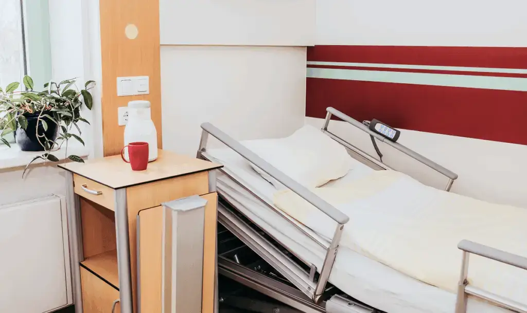 Krankenhausbett neben einem Nachttisch mit roter Tasse und weißer Kanne, am Fenster mit einer grünen Pflanze.