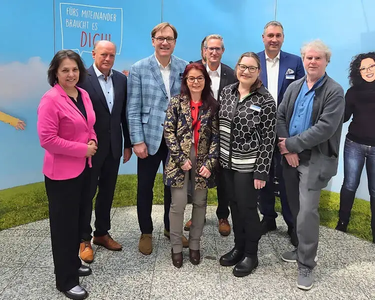 Acht Personen stehen zusammen vor einer Wand mit der Aufschrift ‚Fürs Miteinander braucht es dich‘ und lächeln in die Kamera.