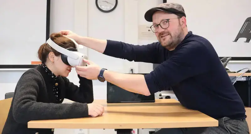 Ein Mann setzt einer sitzenden Frau eine Virtual-Reality-Brille auf, beide befinden sich in einem Seminarraum.