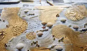 Reliefbild in Sand- und Goldtönen mit Wellenlinien und Muscheln; zeigt ein kreatives Ergotherapie‑Projekt auf einem Arbeitstisch.