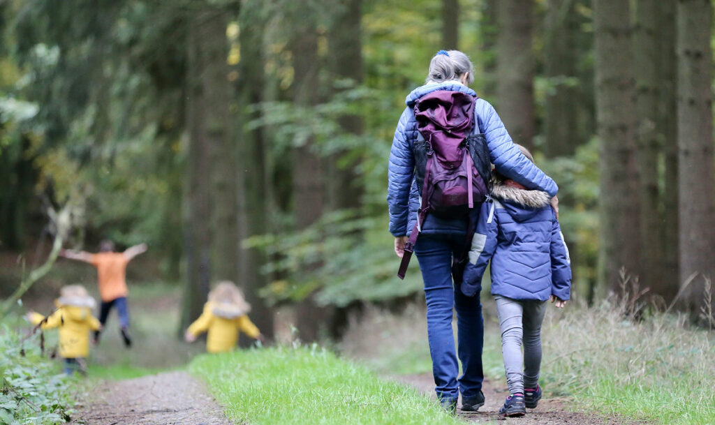 Erwachsene Person geht Arm in Arm mit einem Kind auf einem Waldweg, während weiter vorne drei weitere Kinder spielend auf dem Weg unterwegs sind
