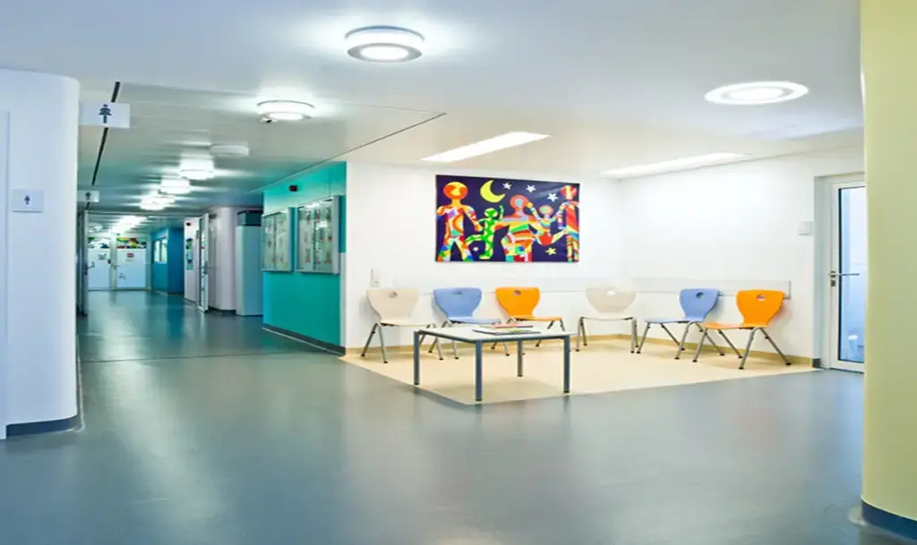 Heller Wartebereich eines Krankenhauses oder einer Arztpraxis mit bunten Stühlen um einen niedrigen Tisch, farbenfrohem Kinderbild an der Wand und einem langen, gut beleuchteten Flur im Hintergrund.