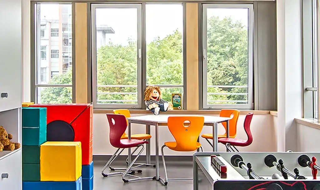 Heller Spiel- und Aufenthaltsraum mit großem Fenster, bunten Kinderstühlen um einen Tisch, einer Puppe darauf und einem Tischkicker im Vordergrund.