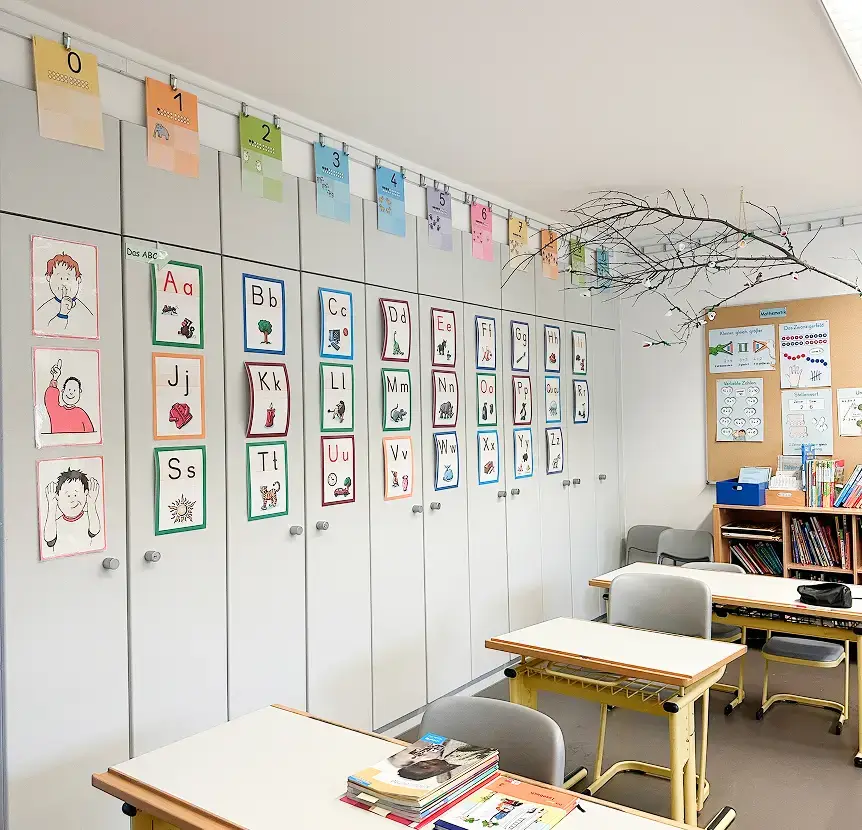 Heller Klassenraum mit kindgerecht gestalteten Alphabet-Postern an einer Schrankwand, farbigen Zahlenkarten oben, mehreren Schultischen und Stühlen sowie einem Bücherregal und Lernplakaten im Hintergrund.