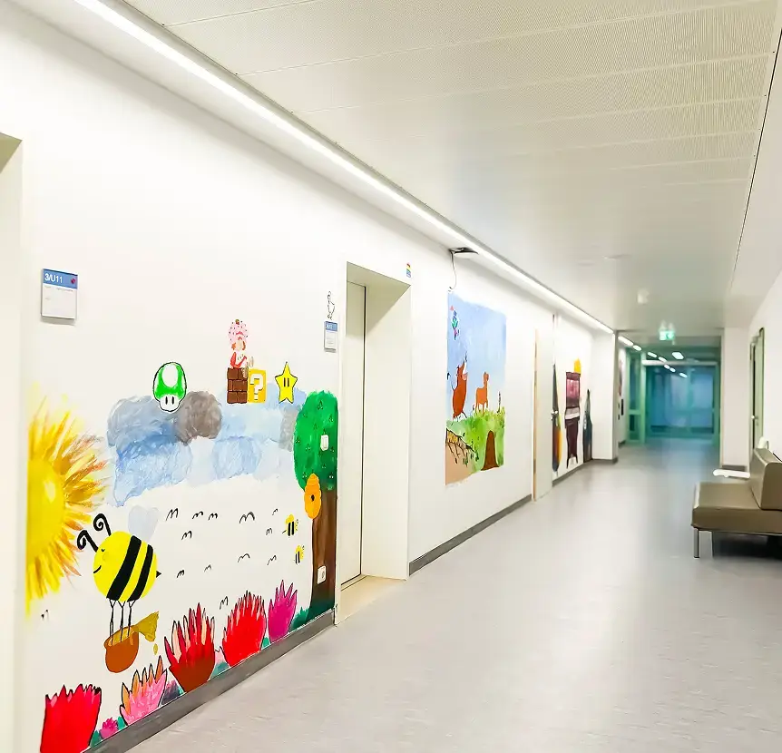 Heller Krankenhausflur mit farbenfrohen Kinderzeichnungen an den Wänden, rechts Sitzbänke und am Ende des Gangs eine Glastür.