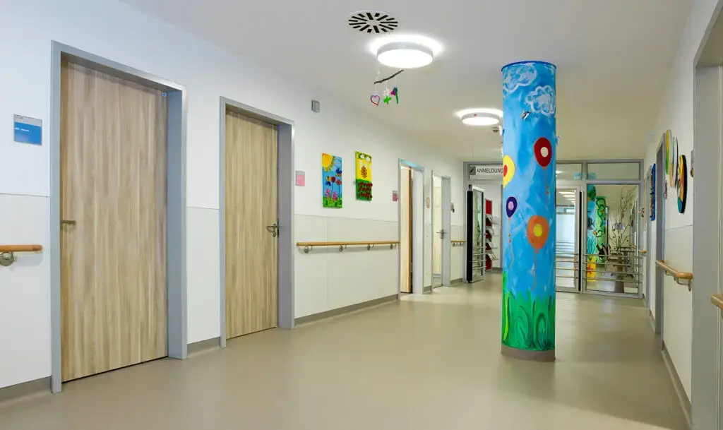 Heller Klinikflur mit Holztüren, bunten Kinderbildern an den Wänden und einer farbenfroh bemalten mittleren Säule mit Luftballons und Wolkenmotiven.