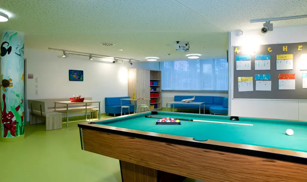 Freizeitraum mit Billardtisch im Vordergrund, blauen Sofas und Sitznischen im Hintergrund, einer bunt bemalten Säule sowie Pinnwand und Spielen in einer hellen Klinikumgebung