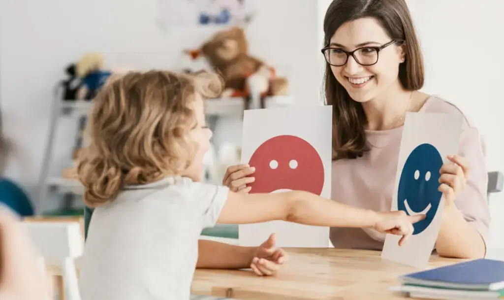 Eine junge Frau mit Brille, die einem Kind einen lachenden und einen traurigen Smiley zeigt, das Kind deutet mir ihrer Hand fröhlich auf den lachenden Smiley
