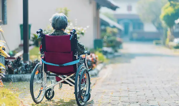 Ältere Person sitzt in einem Rollstuhl und fährt auf einem gepfalsterten Weg.